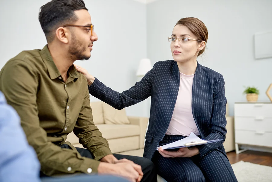 Therapist in a pinstriped suit comforting a client by placing a hand on his shoulder in a cozy office setting.