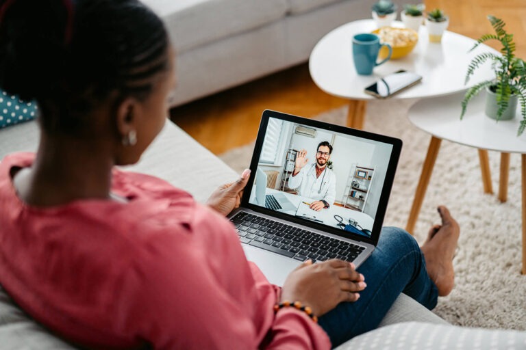 Woman at home on a laptop video calling a doctor during a telemedicine appointment in a cozy living room.