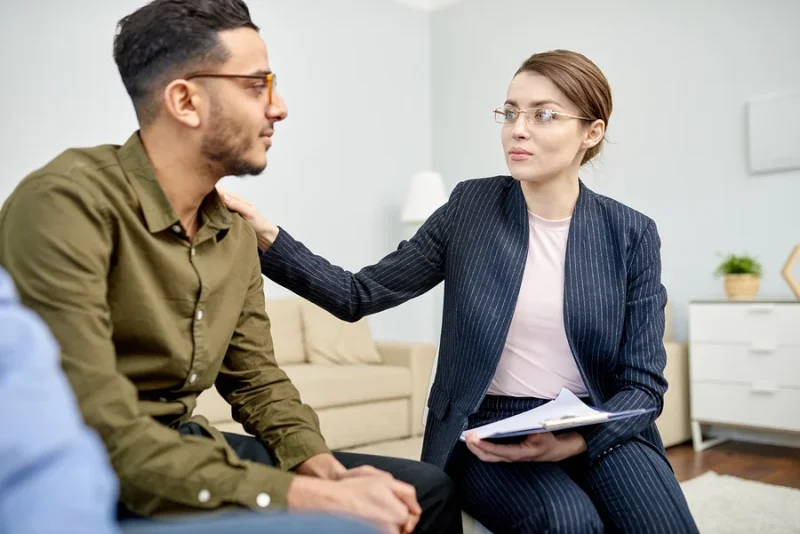 Therapist in a pinstriped suit comforting a client by placing a hand on his shoulder in a cozy office setting.